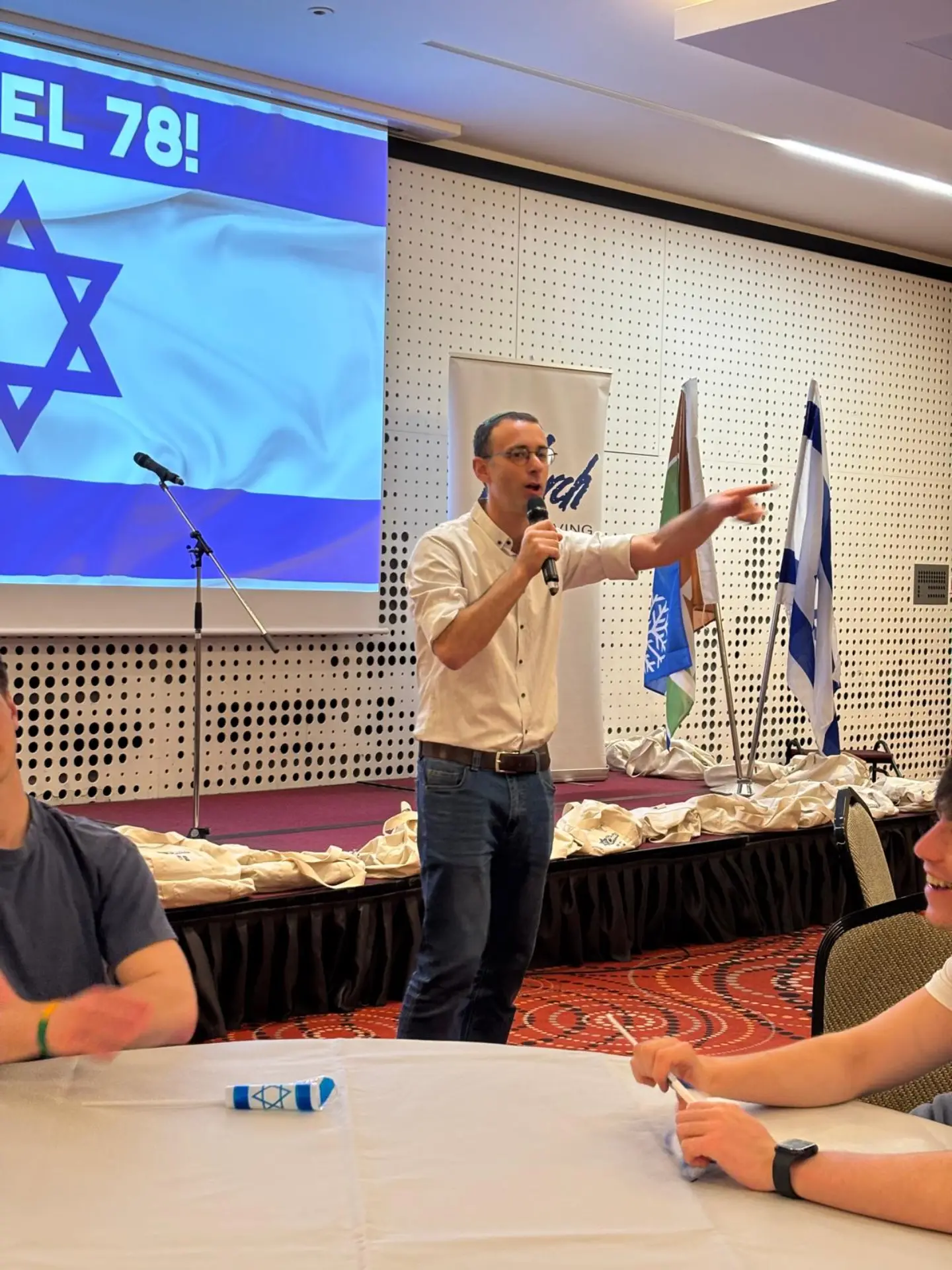יואל פרנקנבורג מרצה מול בני נוער באולם עם דגלי ישראל